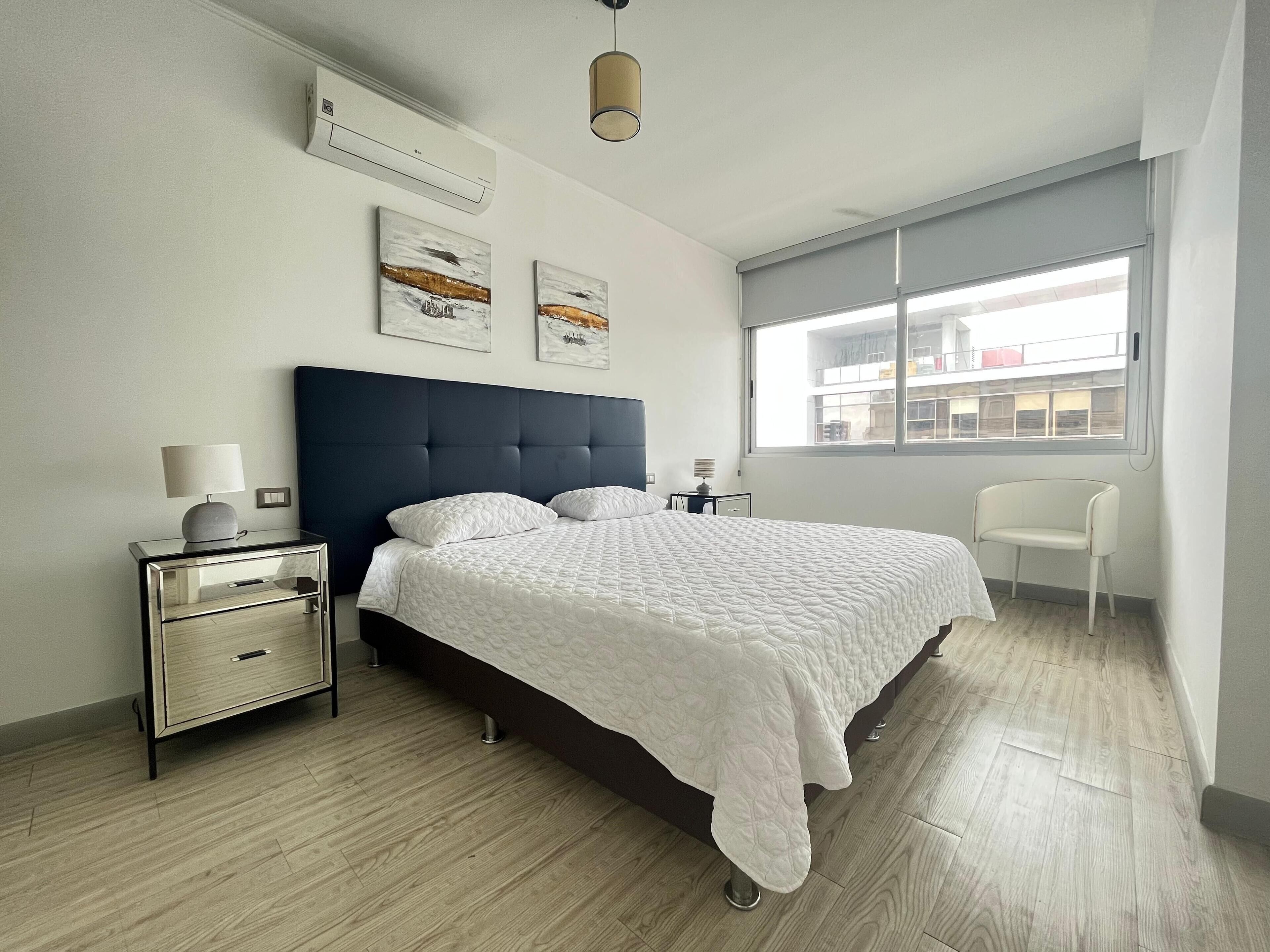 A modern bedroom with a large bed, a bedside table, a chair, and a window with a view of buildings.