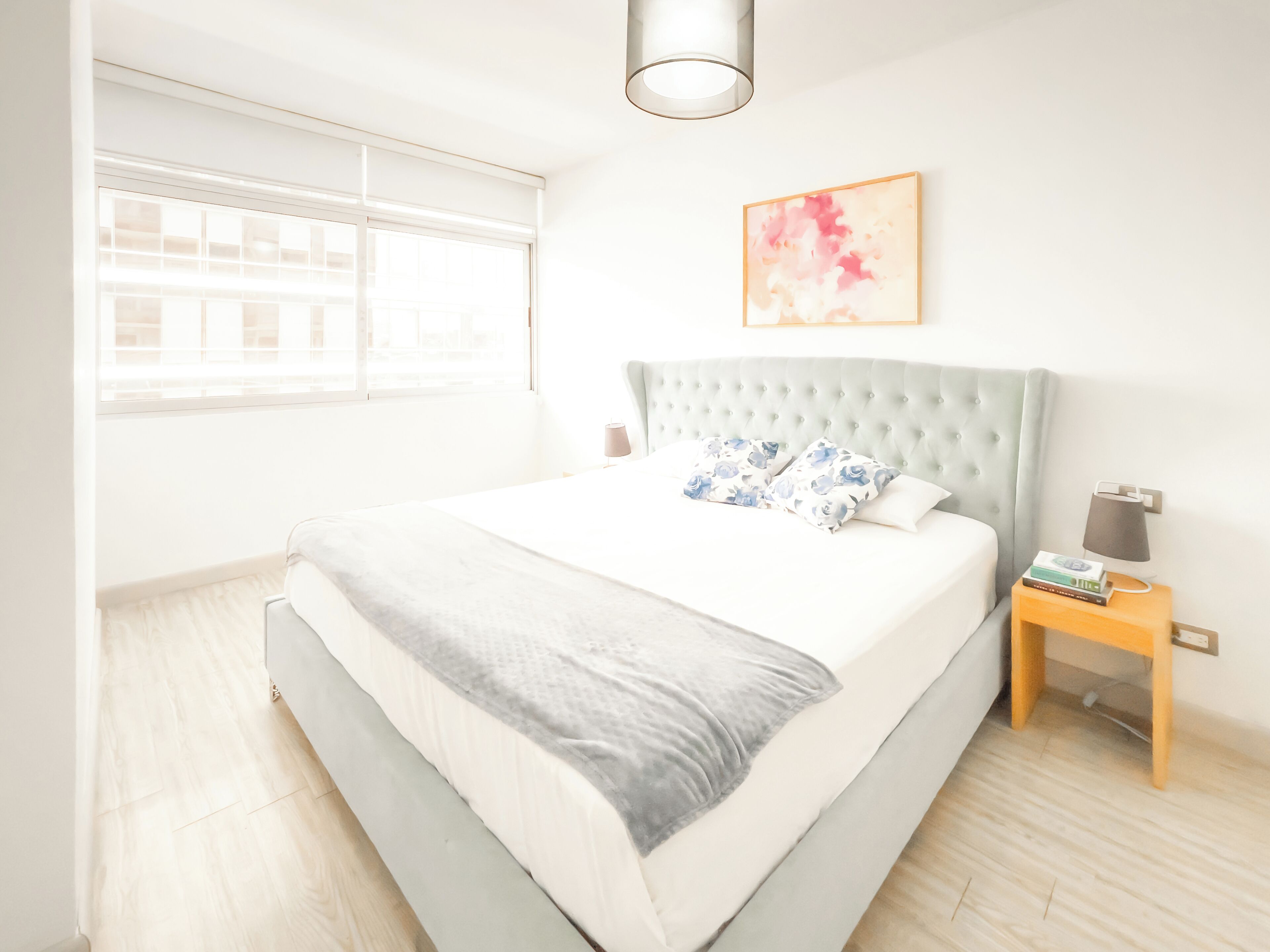 A modern bedroom with a large bed, a tufted headboard, a bedside table with a lamp, and a window with blinds.