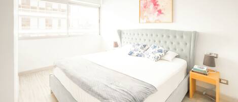 A modern bedroom with a large bed, a tufted headboard, a bedside table with a lamp, and a window with blinds.