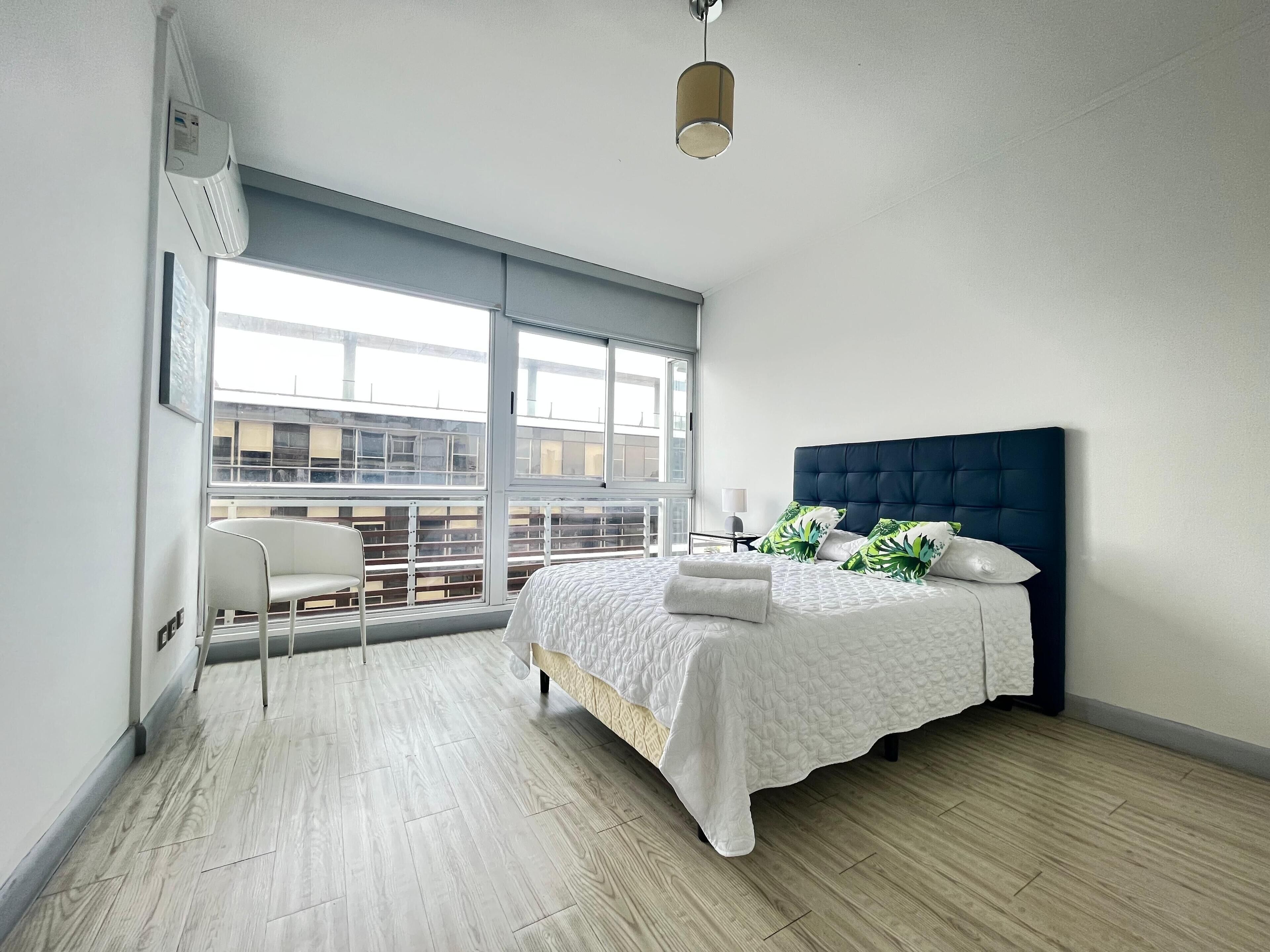 A modern bedroom with a large window, a bed with a dark headboard, and a white chair.