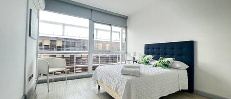 A modern bedroom with a large window, a bed with a dark headboard, and a white chair.