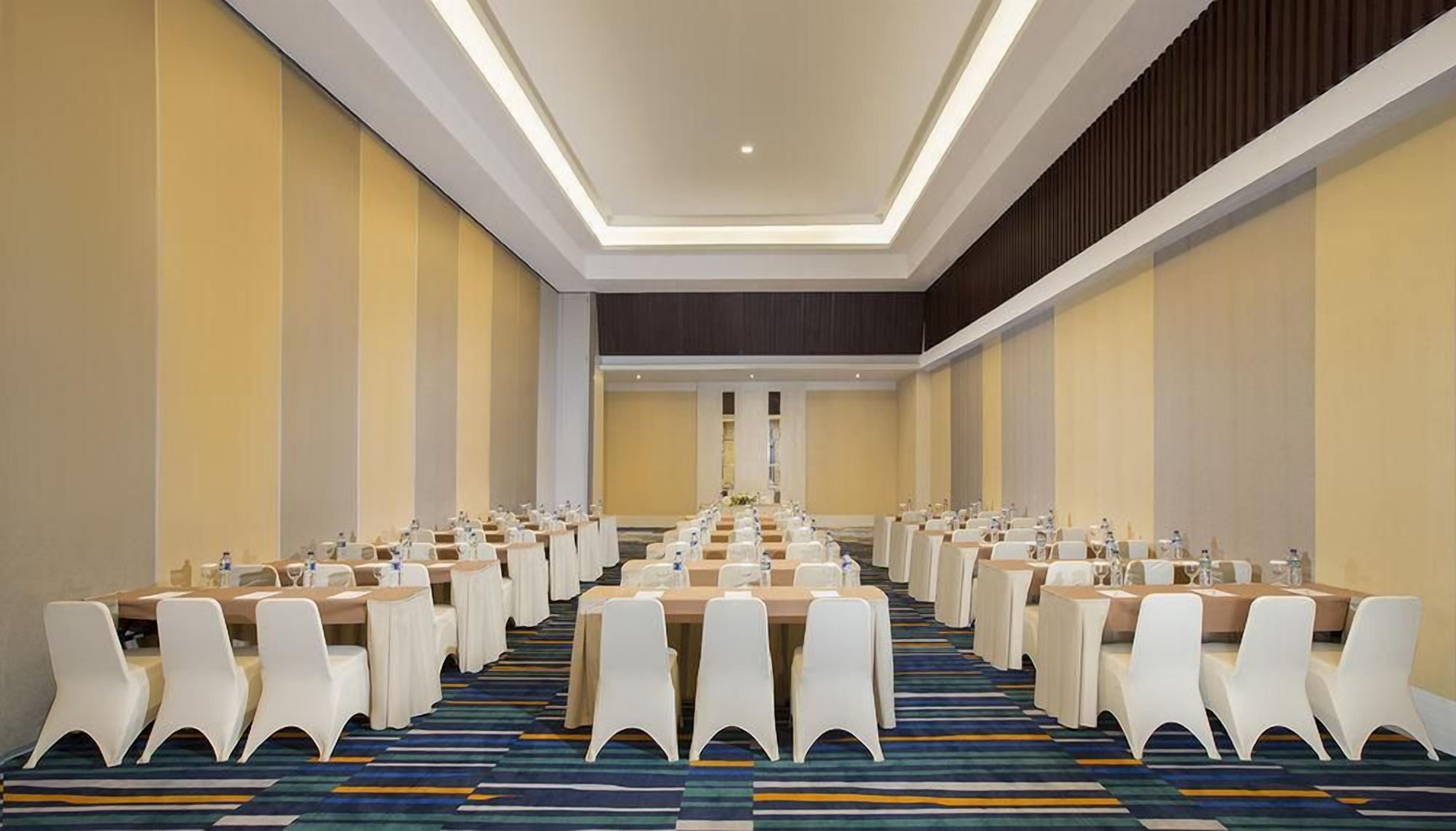 Un salón de conferencias con filas de mesas y sillas, una alfombra estampada y una paleta de colores neutros.