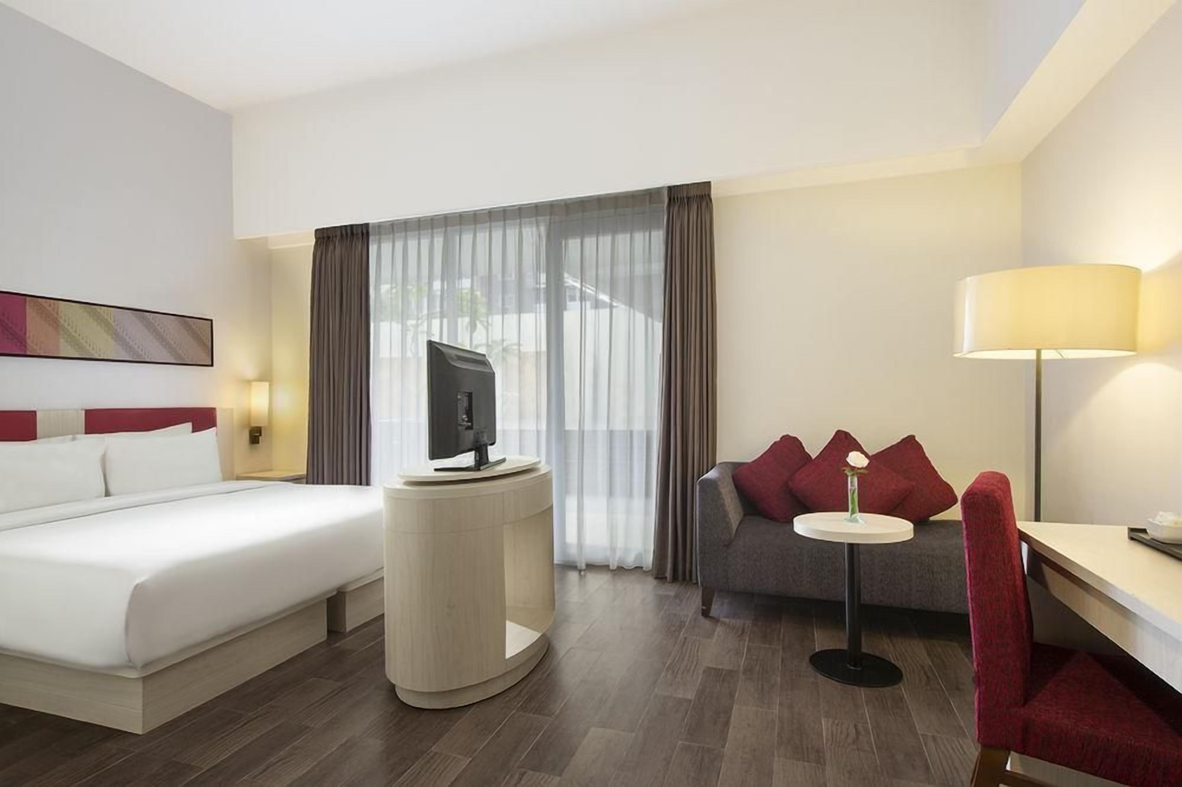 Una habitación de hotel con una cama, un televisor sobre un mueble bajo, un sofá con cojines rojos, una mesita con una lámpara y un ventanal con cortinas.