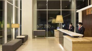 Zona de recepción de un hotel, con un mostrador, dos áreas de espera con sofás y un ventanal que ofrece vistas del interior.