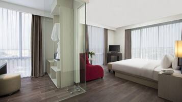 A hotel room with a bed, a TV, a desk, a chair, and a glass partition.