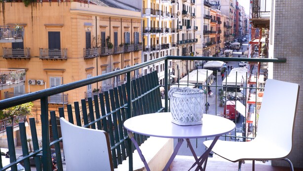 A balcony with a round table and two white chairs, overlooking a street with buildings and parked vehicles. - Porta Amari (Palermo)
