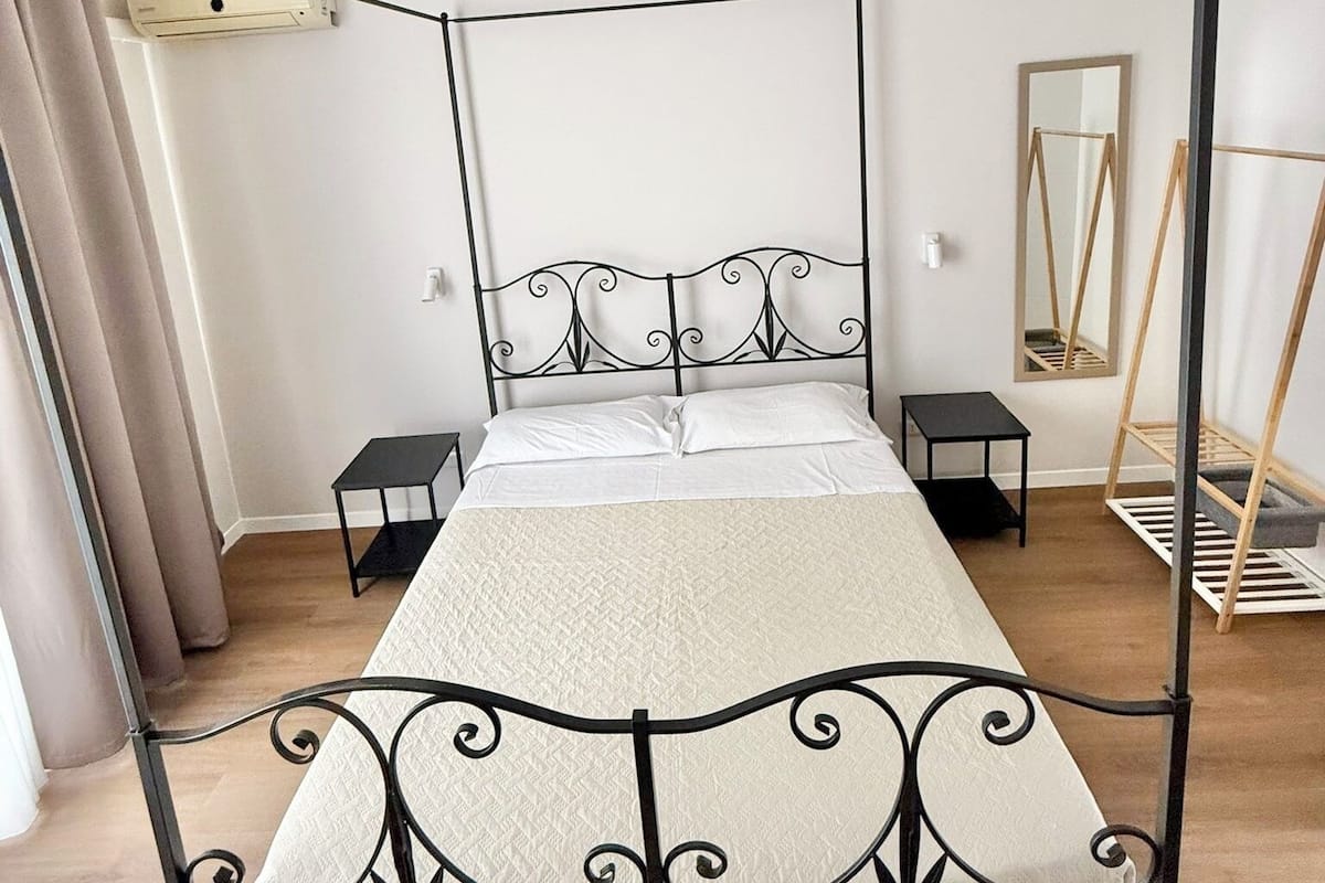 Una cama con dosel, con una colcha y almohadas blancas, una mesita de noche pequeña, un aire acondicionado de pared y un espejo en la pared.