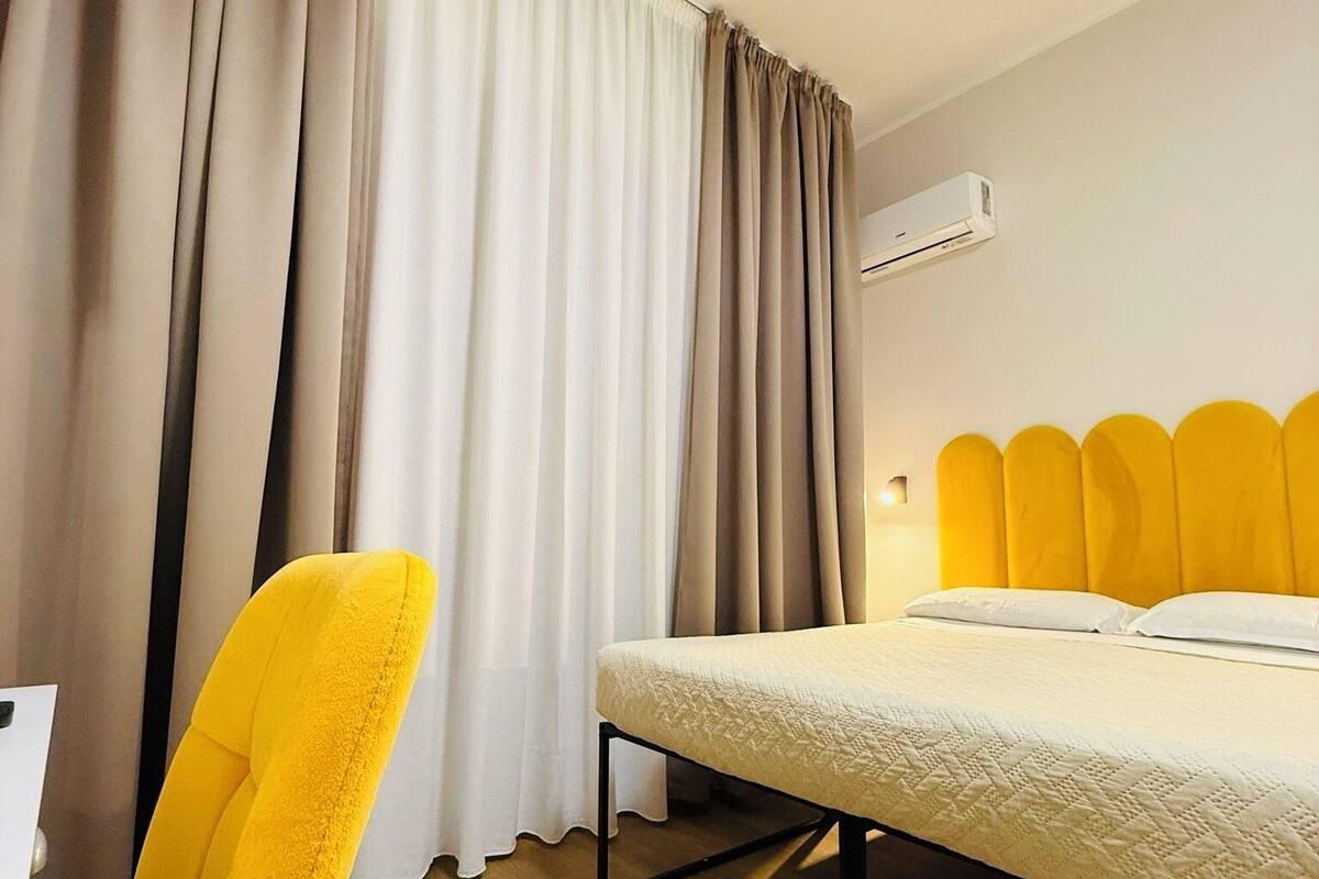 Una habitación de hotel con cama, una silla, cortinas y un equipo de aire acondicionado.