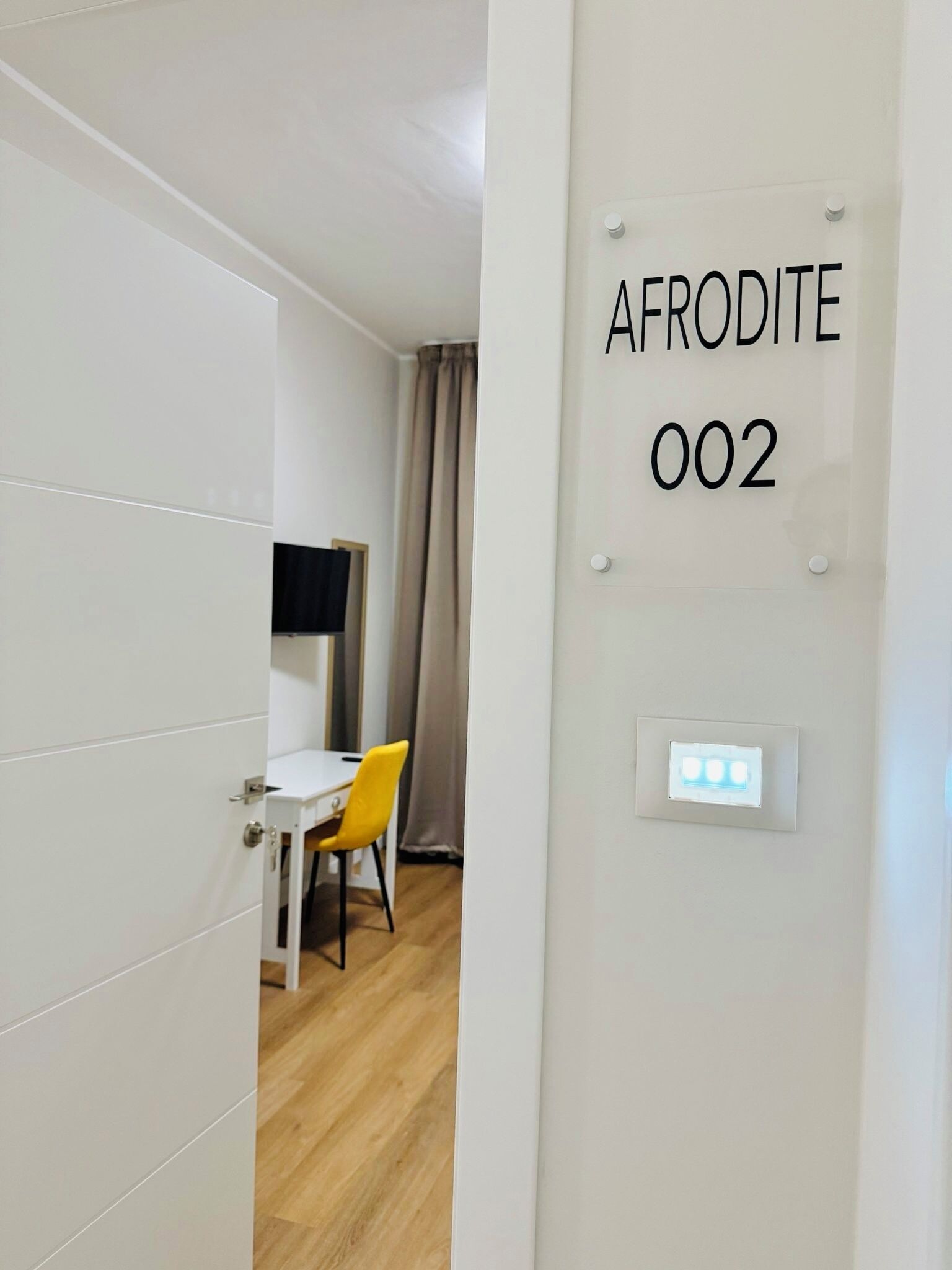 A hotel room with a white door, a bed, a small table with a chair, a television, and a wall-mounted light switch.