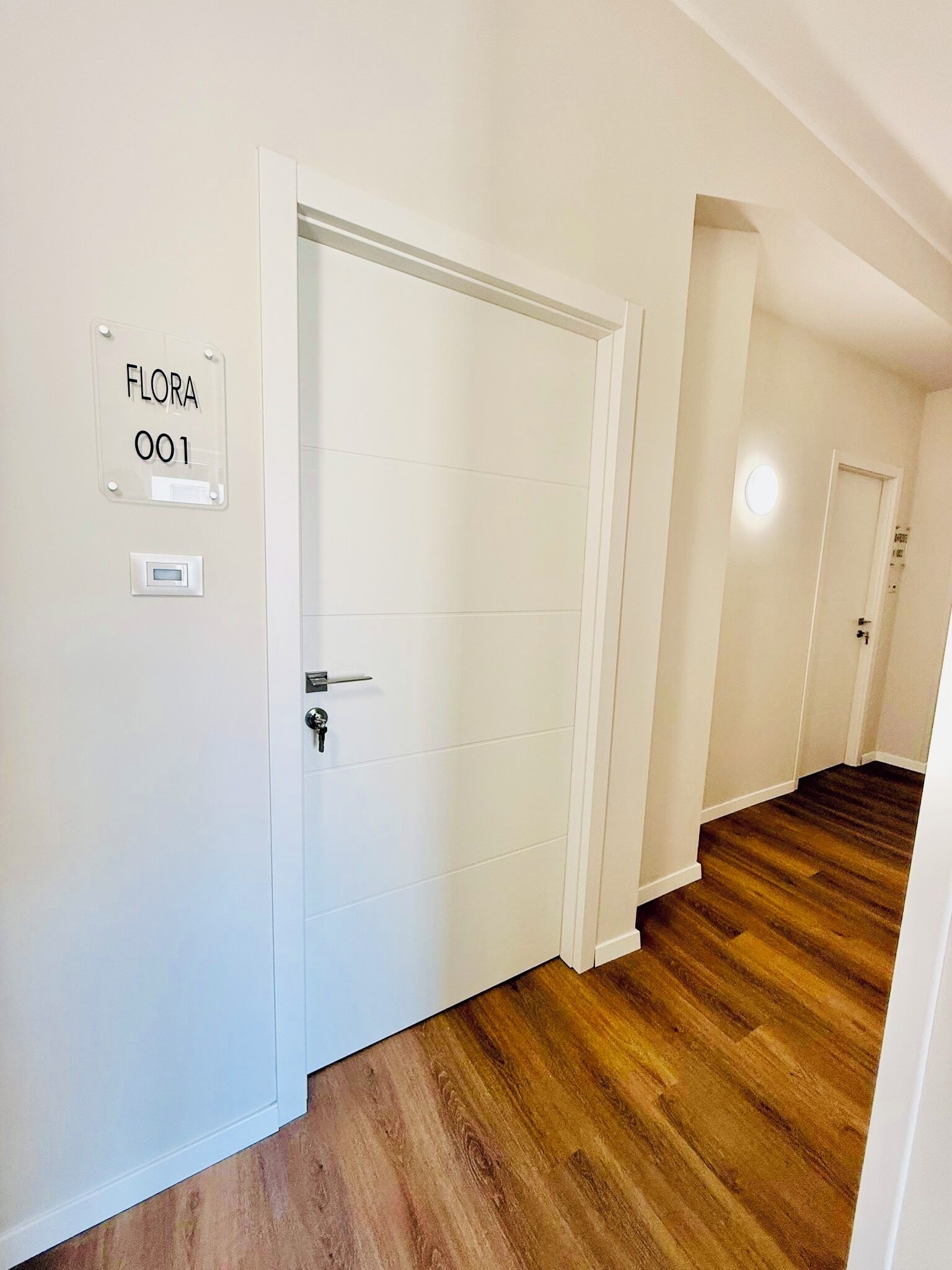 A hallway with a white door labeled 'FLORA 001' and a thermostat on the wall.