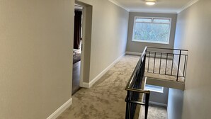 A narrow hallway with beige walls, a carpeted floor, a window with a view of trees, and a black metal railing on the right. - Luxurious Living in the Heart of Bromley (Bromley)
