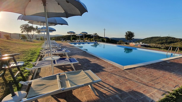 Una piscina con sillas de descanso y sombrillas, rodeada por un patio de ladrillo y vegetación.