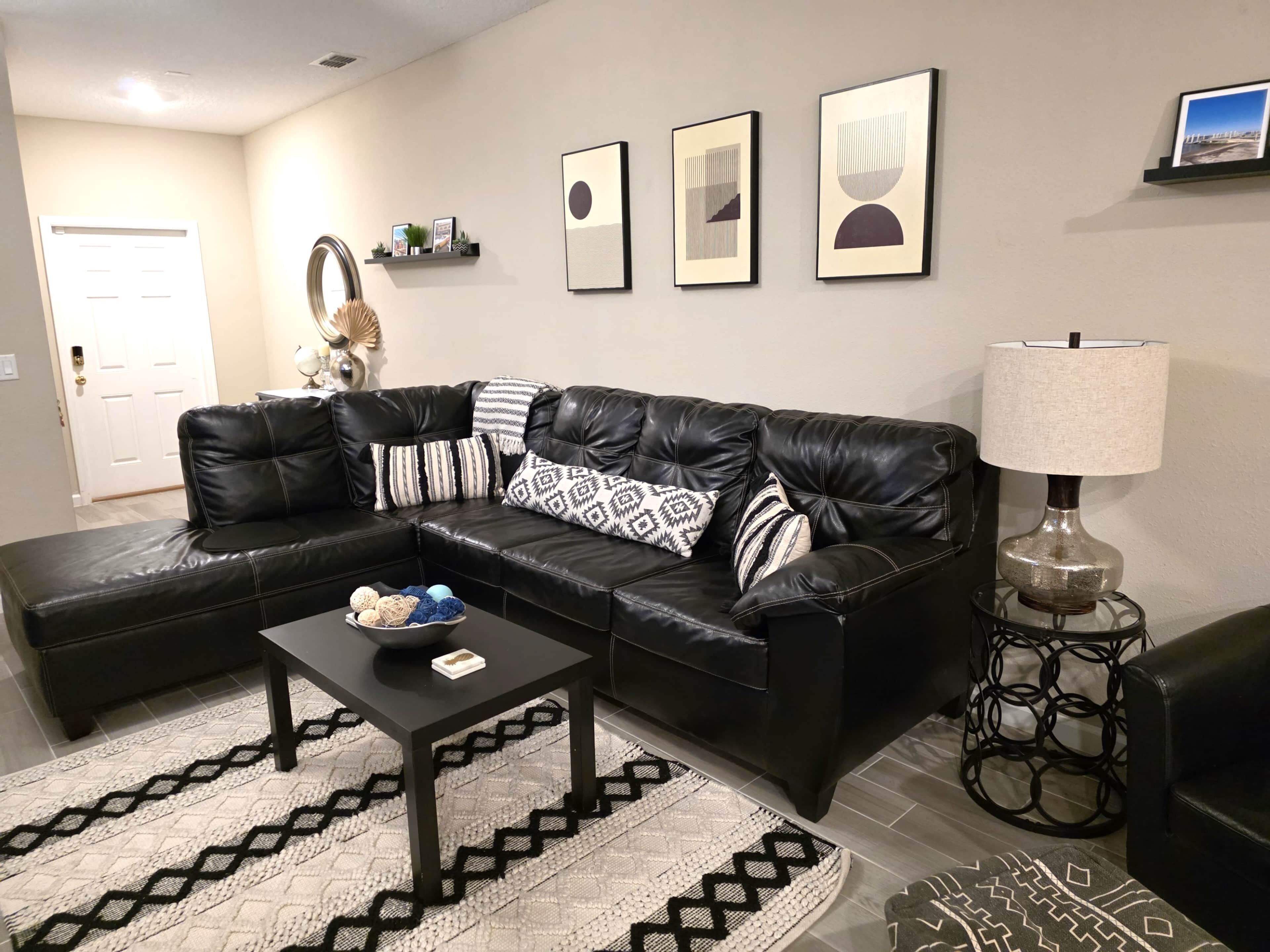 A black leather sectional sofa, a small black coffee table, a patterned rug, a lamp, and framed artwork on the wall.