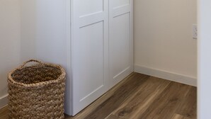 Um guarda-roupa branco com duas portas e puxador, uma cesta de vime e uma janela com persianas.
