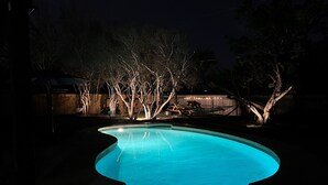 Une piscine éclairée la nuit, entourée d’arbres et bordée d’une clôture en bois.