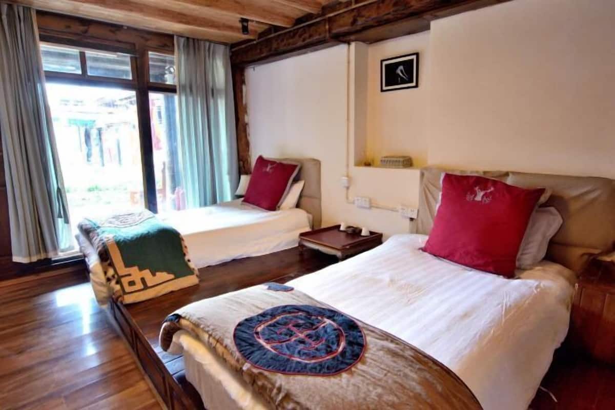 A room with two beds, wooden flooring, a window with curtains, a wall-mounted light, and a small table.