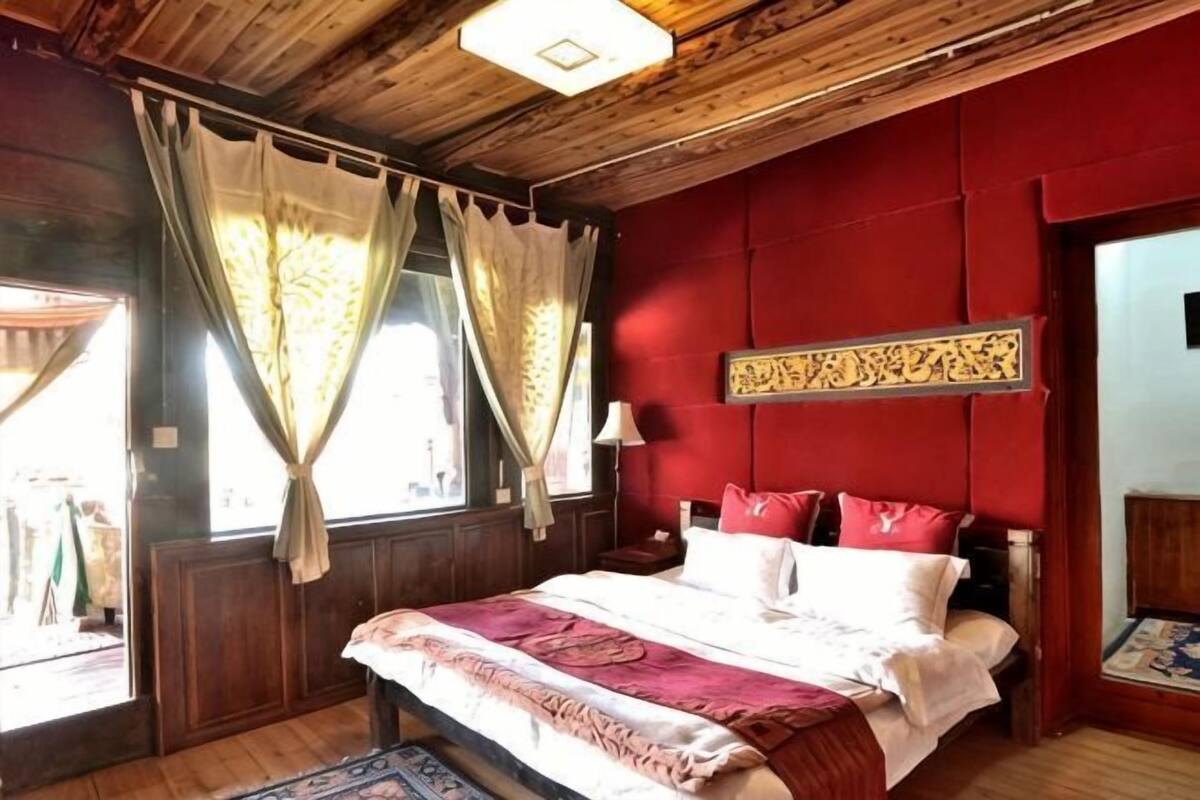 A room with a wooden ceiling, a bed with a red and white bedspread, a red wall with a decorative panel, and a window with curtains.