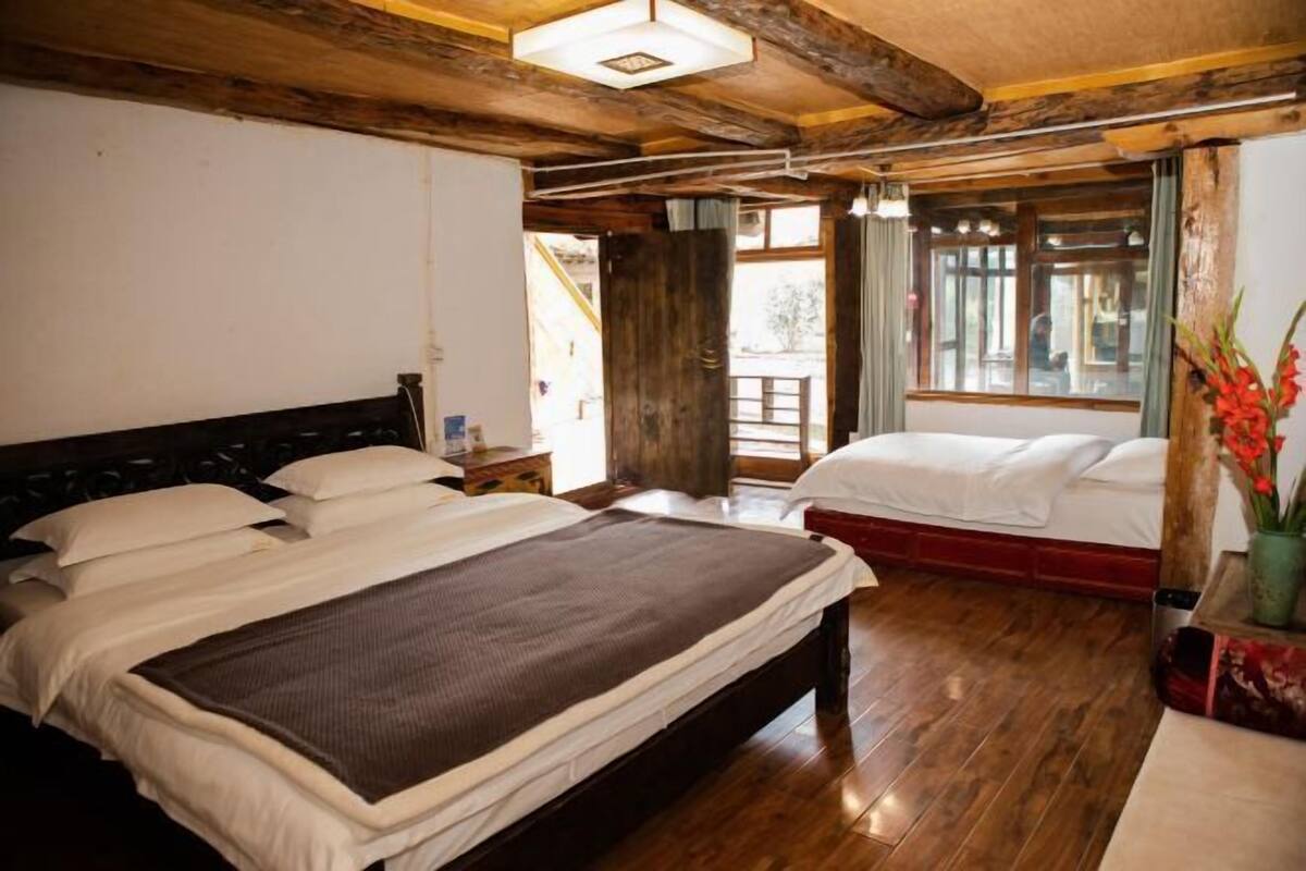 A room with two beds, wooden flooring, a large window, and a wooden ceiling.