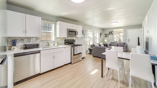 A modern kitchen with white cabinets, stainless steel appliances, and a dining area with white chairs. - Convenient Tulsa Home - 2 Mi to Downtown! (Tulsa)