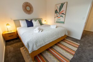 Una cama bien tendida con una manta blanca y almohadas, una lámpara de noche, una obra de arte en la pared y una alfombra estampada.