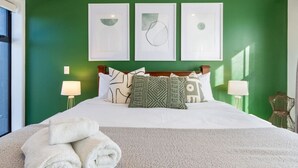 Una cama bien hecha con sábanas blancas y un plaid beige, una pared verde con tres obras de arte enmarcadas y dos lámparas de noche.