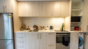 Una cocina compacta con armarios de madera clara, un refrigerador de acero inoxidable, un horno empotrado y una lavadora.