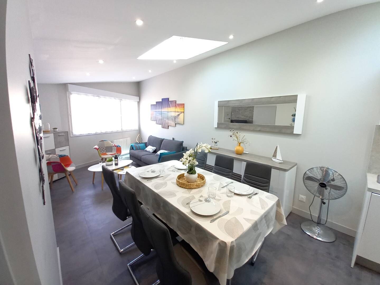 Uma sala de jantar moderna com uma mesa posta para quatro pessoas, um sofá e um ventilador no canto.