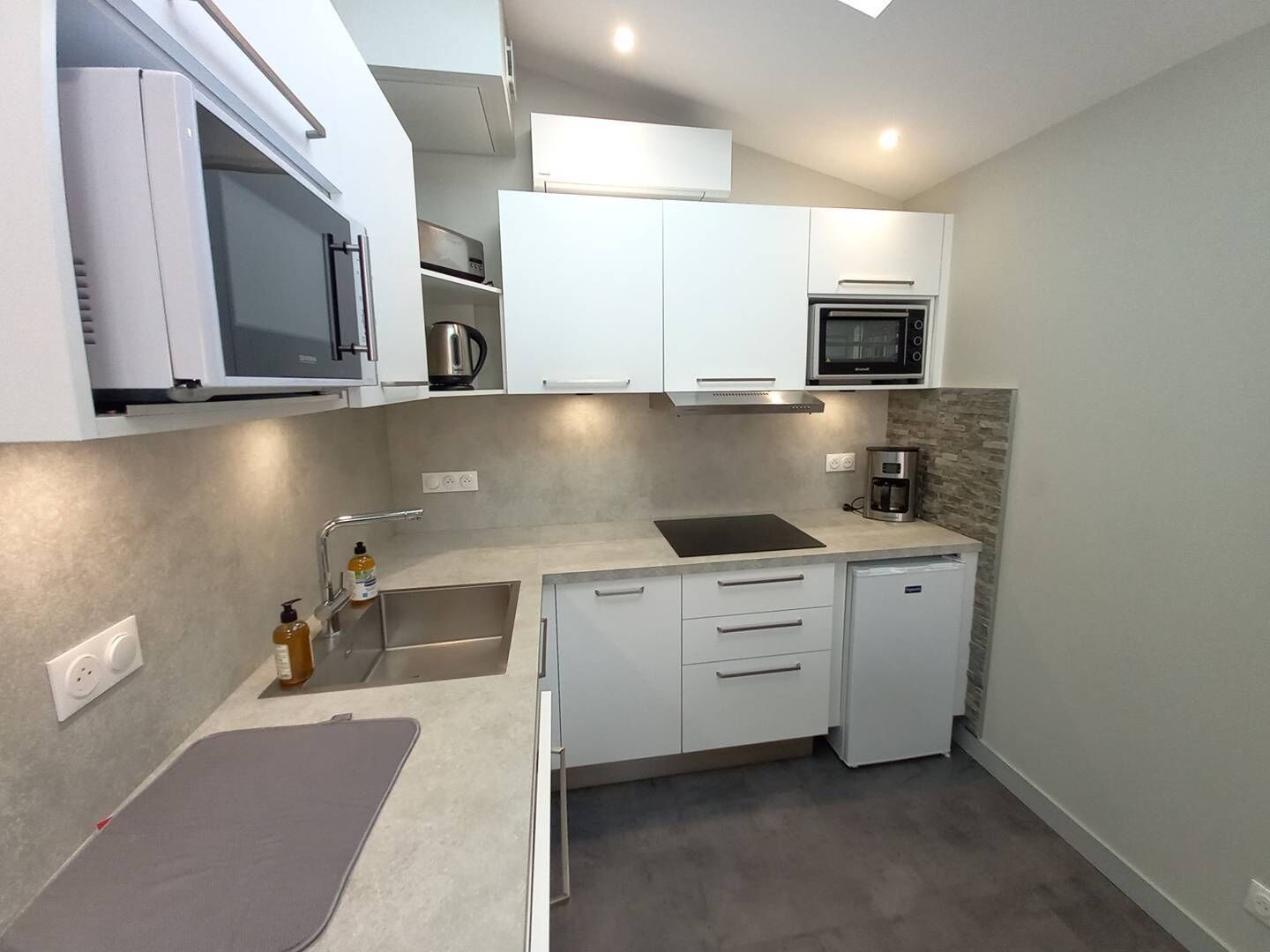 Una cocina compacta con armarios blancos, microondas integrado, cafetera y lavamanos.