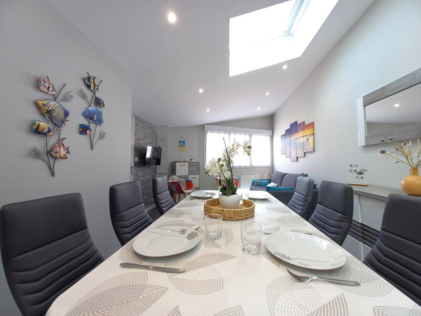 Een moderne eetruimte met een hoge plafond, een dakraam en een lange tafel gedekt voor vier personen.