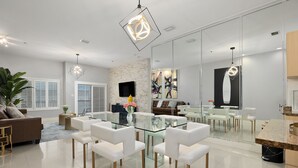 A modern living room with a glass dining table, white chairs, a sofa, and a large mirror. - Elegant Condo with Bay Views, Pool & Hot Tub - Walk to Beach! (South Padre Island)