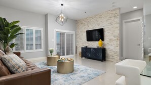 A modern living room with a brown leather sofa, a glass coffee table, and a blue patterned rug. There is a black entertainment unit with a flat-screen TV and a yellow vase. - Elegant Condo with Bay Views, Pool & Hot Tub - Walk to Beach! (South Padre Island)
