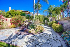 A garden pathway with stone tiles, surrounded by various plants and palm trees.
