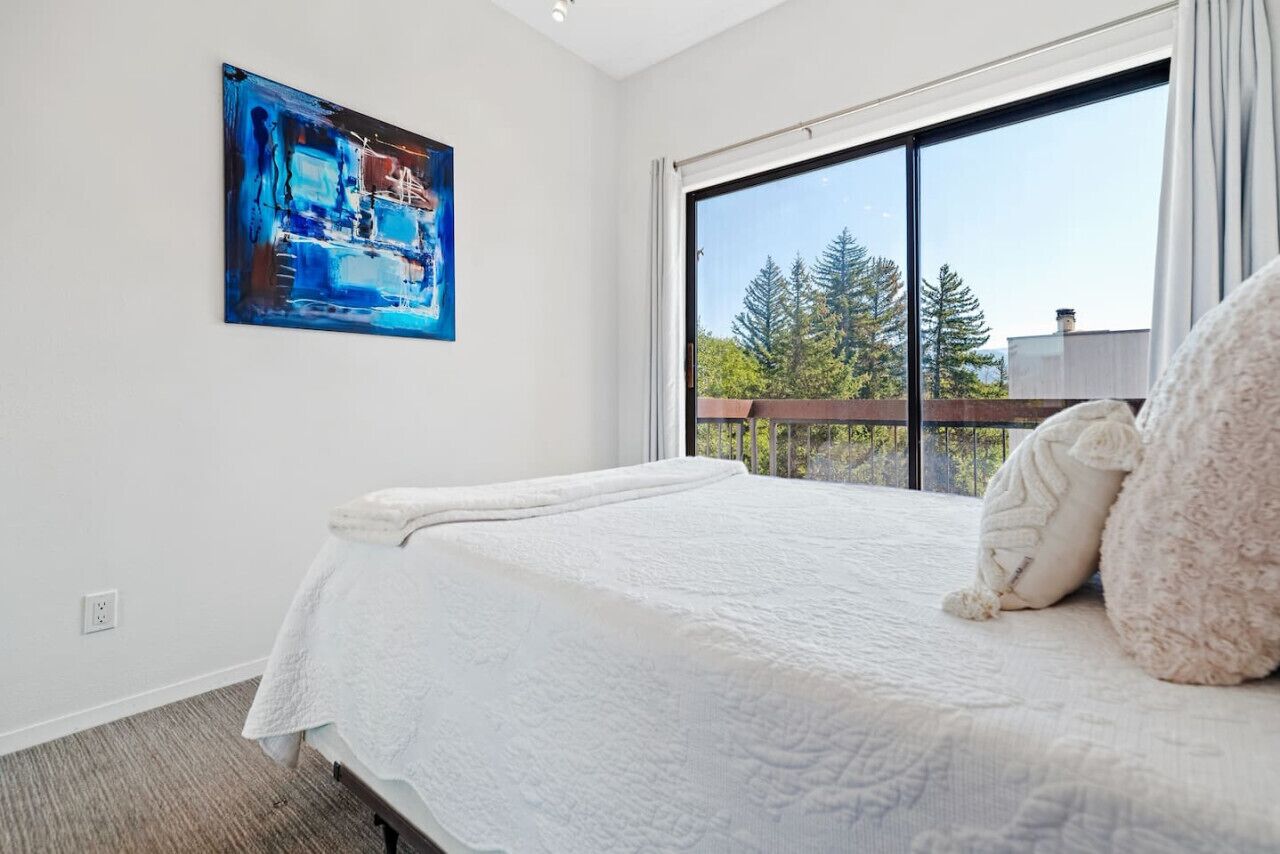 Une chambre avec une grande fenêtre, un lit recouvert d’une couette blanche et un tableau accroché au mur.