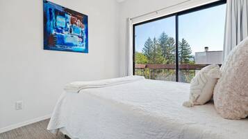 Une chambre avec une grande fenêtre, un lit recouvert d’une couette blanche et un tableau accroché au mur.