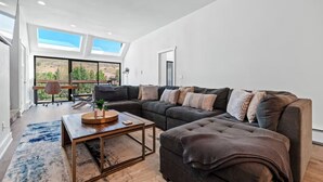 A modern living room with a sectional sofa, a coffee table, and a view of a mountainous landscape through large windows. - Cozy 3BD/2BA Mt. Condo Overlooking Eagle River (Avon)