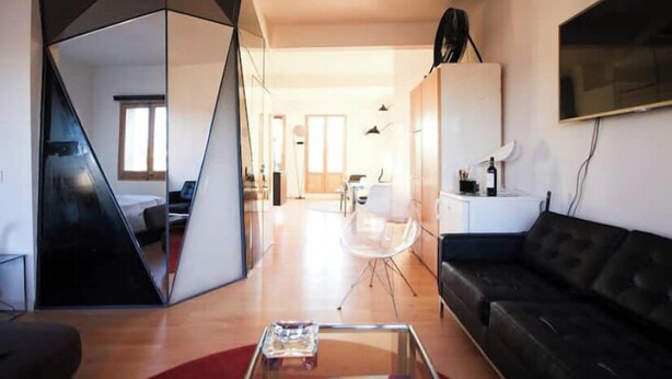 A modern living room with a black sofa, a glass coffee table, and a clear chair. There is a TV mounted on the wall and a window providing natural light. - Diamond Penthouse & Terrace (Barcelona)