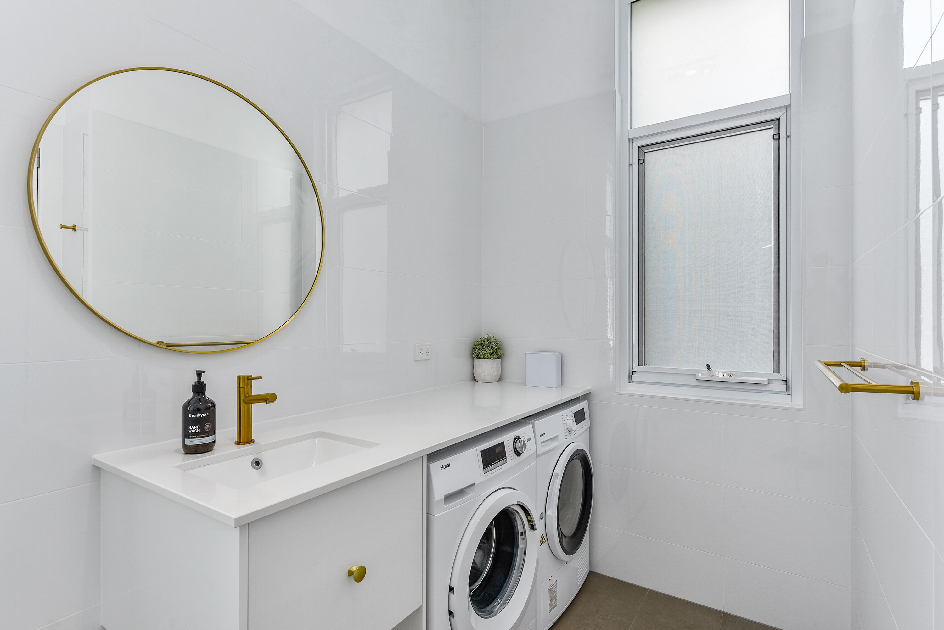 A modern bathroom with a white vanity, gold fixtures, a round mirror, a washing machine, and a window.