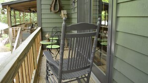 A wooden deck with a chair and a table, a hanging light fixture, and a window with a view of greenery. - Quaint cabin with a creek in front that will lull you to sleep and pets allowed (Hiawassee)