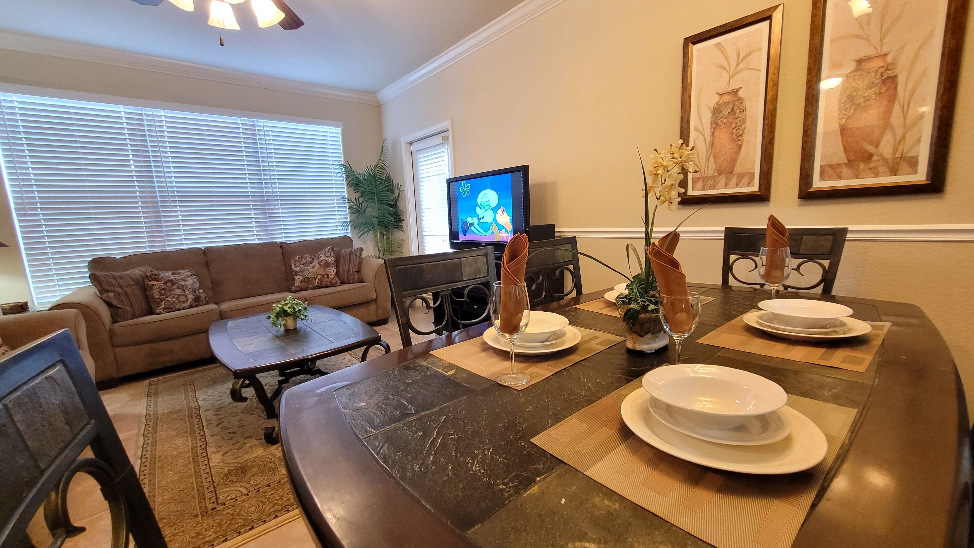 A dining area with a dark wood table set for four, surrounded by chairs. A television is mounted on the wall, and there are framed artworks above the table.