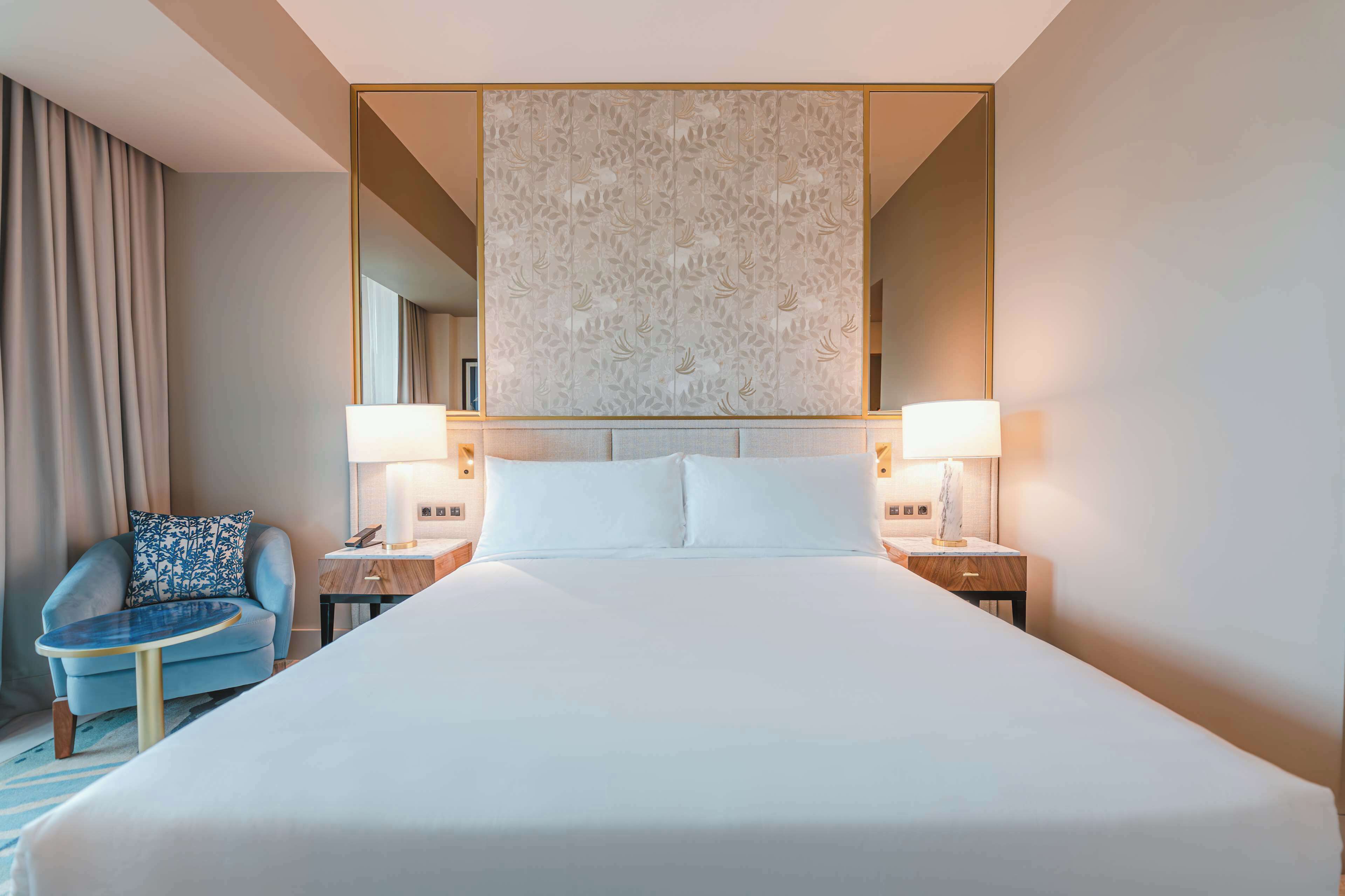 A large bed with white linens, two bedside lamps, a chair with a blue cushion, and a decorative wall panel.