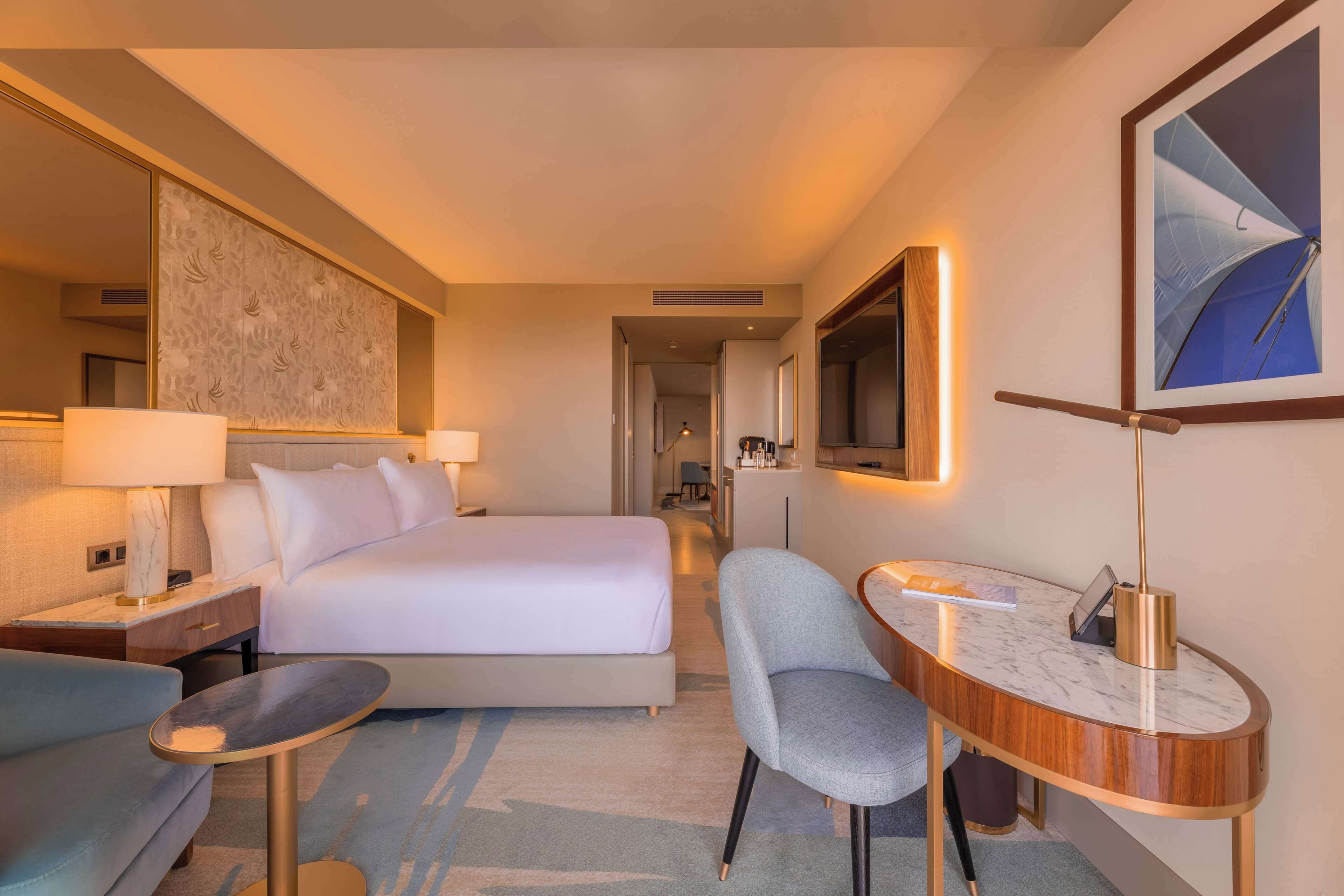 A hotel room with a bed, a desk, a chair, a lamp, a mirror, and a painting on the wall.