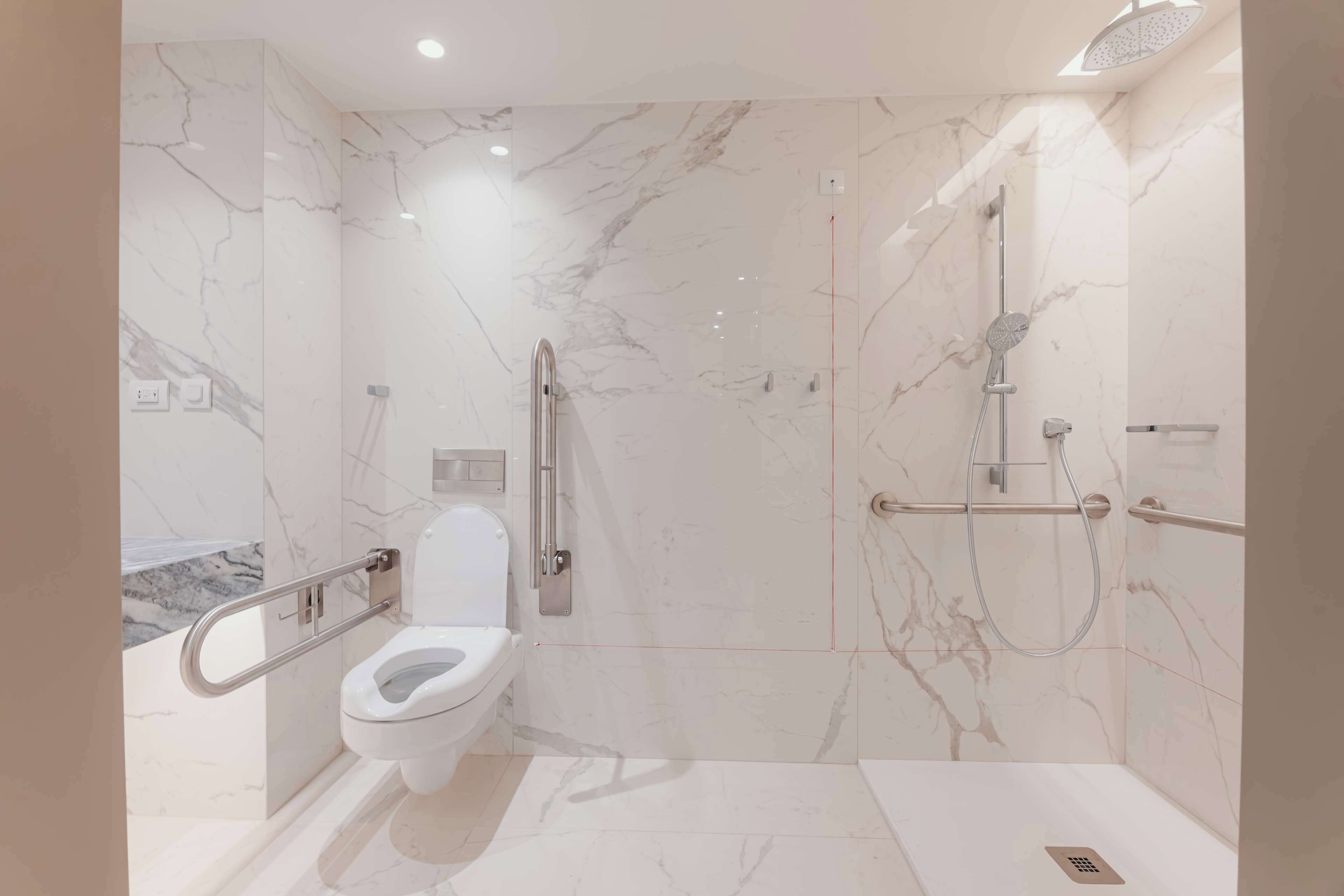 Un baño moderno con suelo de mármol, inodoro blanco, ducha con cabezal de ducha manual y barra de apoyo para mayor seguridad.