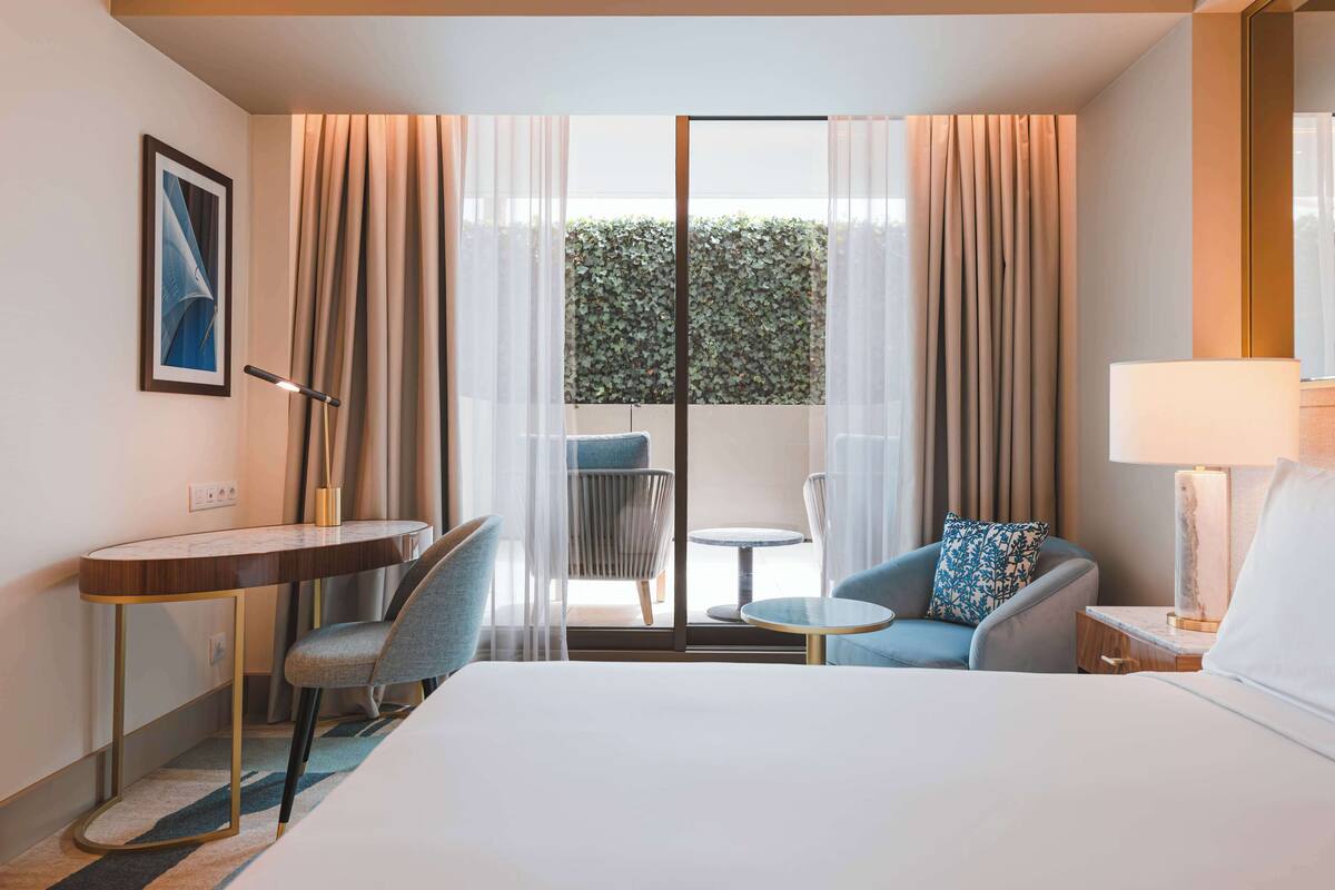 Habitación de hotel con cama, escritorio, dos sillas, una mesita y vista a un seto verde a través de una puerta de vidrio.