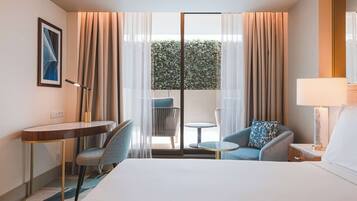 A hotel room with a bed, a desk, two chairs, a small table, and a view of a green hedge through a glass door.