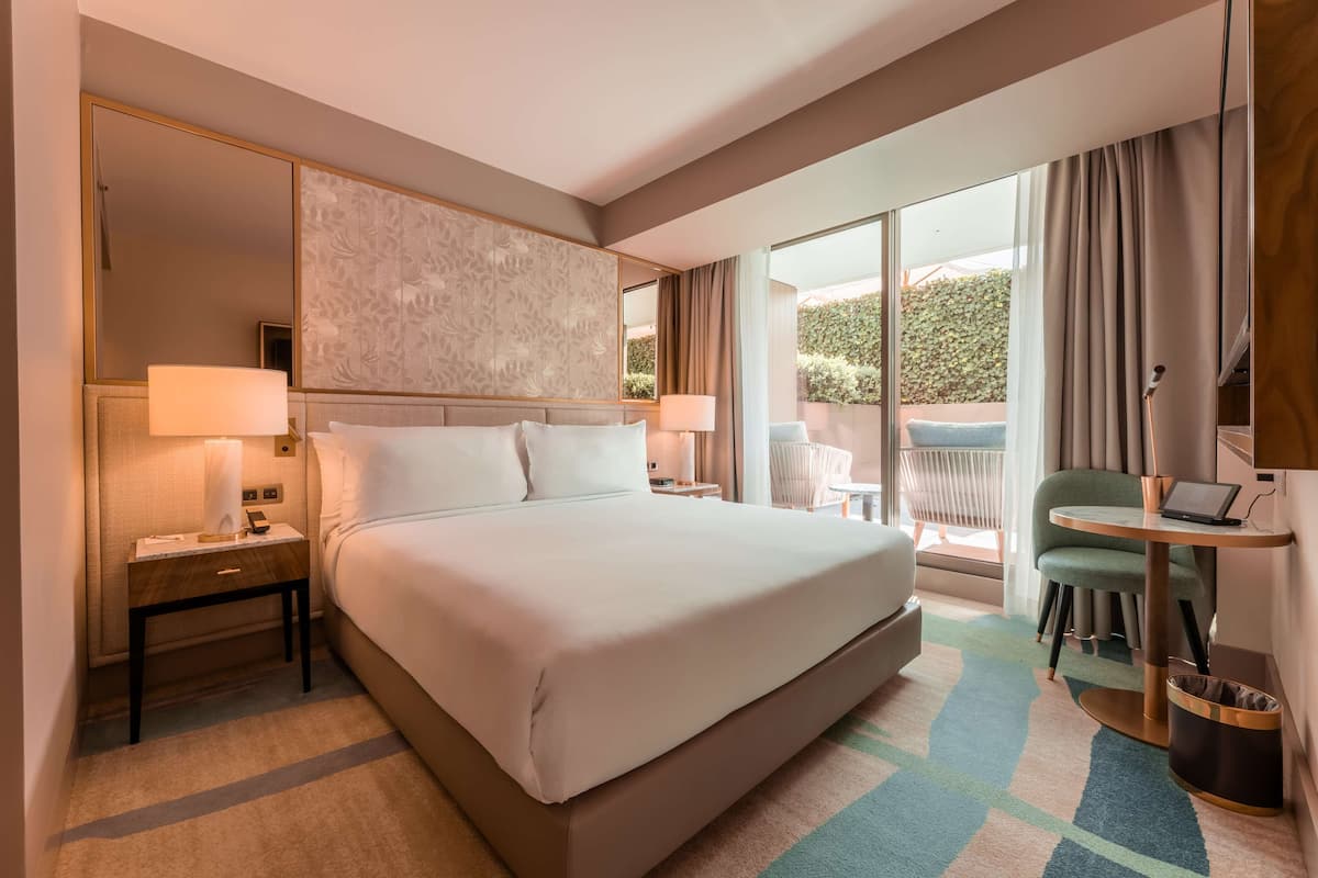 Habitación de hotel con una cama grande, dos mesitas de noche con lámparas, una laptop sobre una pequeña mesa, una silla y vistas al exterior con zonas verdes.