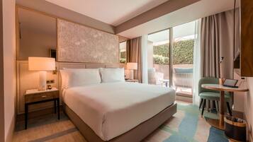 Habitación de hotel con una cama grande, dos mesitas de noche con lámparas, una laptop sobre una pequeña mesa, una silla y vistas al exterior con zonas verdes.