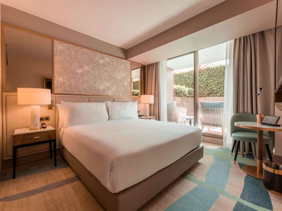Habitación de hotel con una cama grande, dos mesitas de noche con lámparas, una laptop sobre una pequeña mesa, una silla y vistas al exterior con zonas verdes.