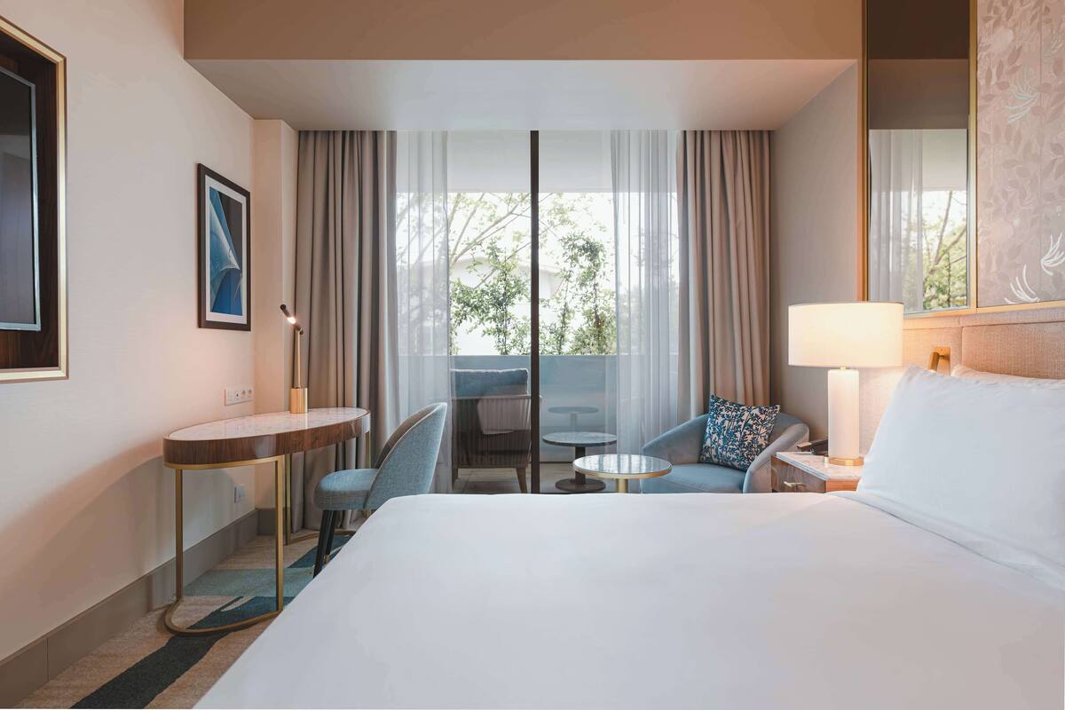 Habitación de hotel con una cama grande, un escritorio, dos sillas, una mesita y un balcón con cortinas.