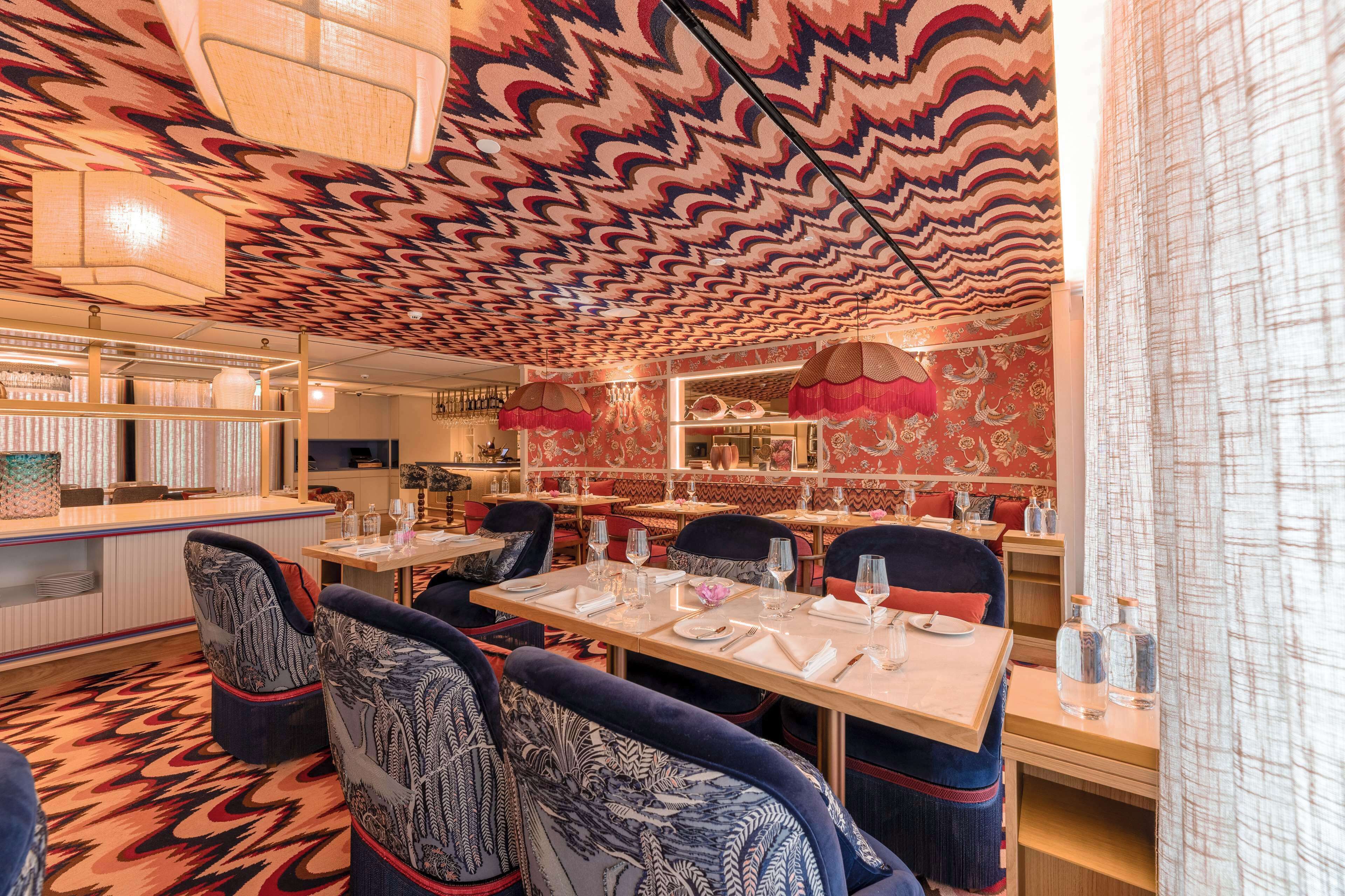 Un restaurante con techo estampado, sillas tapizadas en azul y una zona de bar.