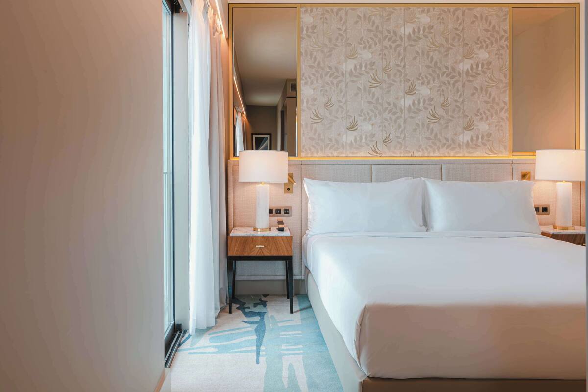Una habitación de hotel con una cama grande, dos mesitas de noche con lámparas, un cabecero decorativo y un espejo amplio.